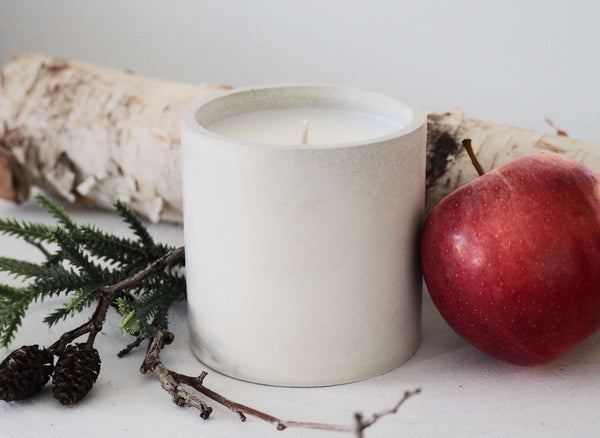 Makana concrete candle on rustic white stool with two fresh red apples and white birch branch in background
